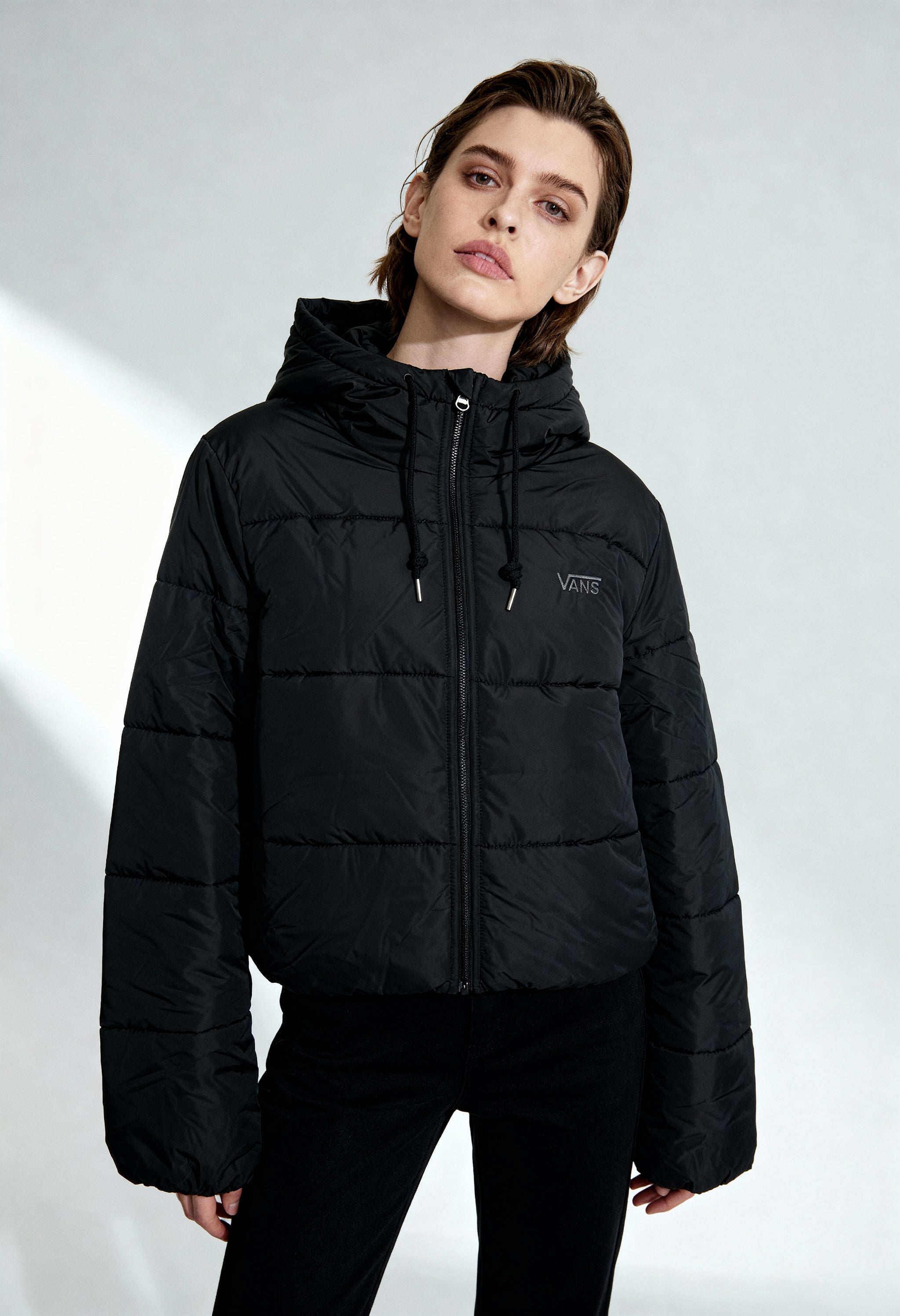 Woman wearing Vans VN0A4B8MBLK1 black puffer bomber jacket with hood and zip fastening, studio shot.
