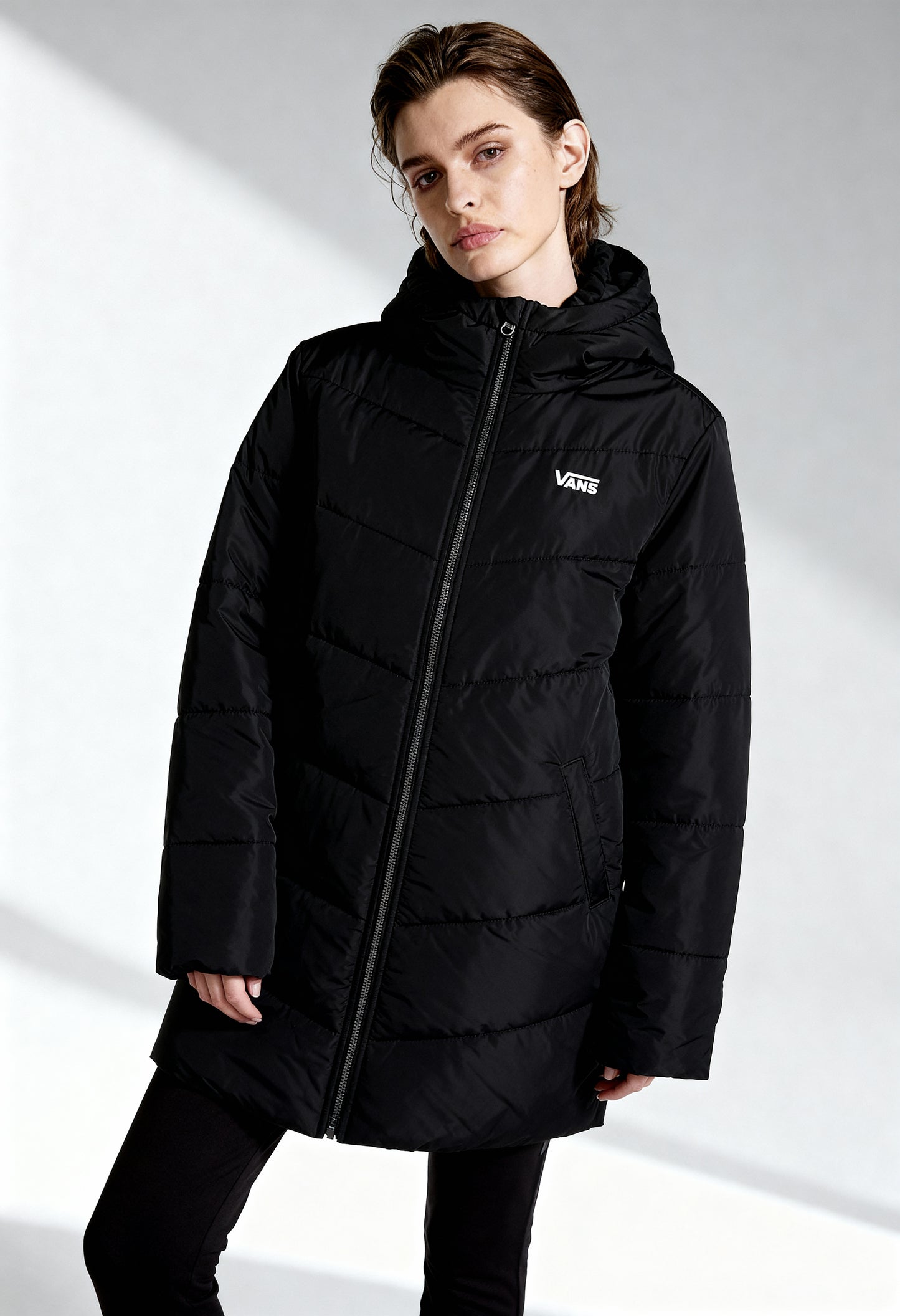 Woman wearing Vans VN0A5JNMBLK1 long black puffer bomber jacket with zip fastening and visible logo, studio shot.
